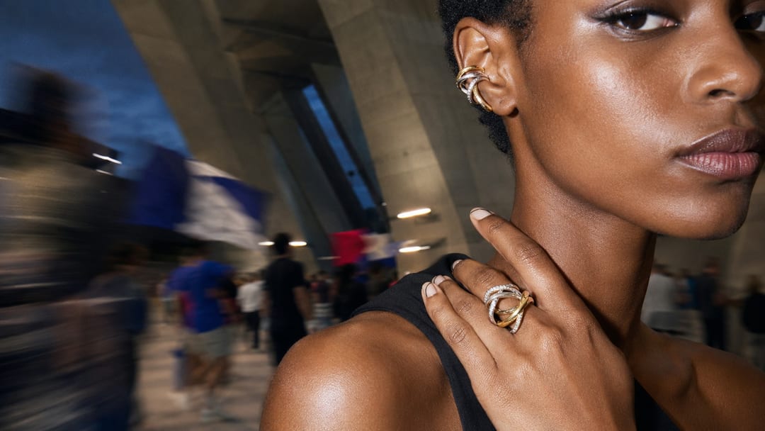 Adult Black model moving through a concrete stadium concourse, wearing a sculptural ear piece and open ring as blurred spectators pass behind her.